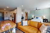 This single-floor unit showcases a thoughtfully designed open-concept layout that seamlessly integrates the kitchen, living, dining, and sleeping areas into one cohesive space. Light wood-look flooring flows throughout, contributing to a clean, contemporary aesthetic.

The kitchen is well-appointed with white shaker cabinetry, granite-style countertops, a subway tile backsplash, stainless steel appliances including a refrigerator, electric range, and microwave, and a single-basin sink with a pull-down faucet. Dark-framed interior doors and gray accents add visual contrast against the neutral walls.

The open living area benefits from large floor-to-ceiling windows and a sliding glass door that invite generous natural light and provide outdoor views. A ceiling fan is centrally positioned over the sleeping area, which is defined by a distinctive paneled accent wall in a muted sage tone.

The bathroom features wood-look flooring, a granite-topped white vanity with ample storage, a large mirror, and direct access to a well-organized walk-in closet with shelving and hanging space. Recessed lighting throughout the unit ensures consistent illumination, reinforcing the space's modern and functional character.