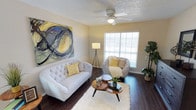 This single-floor home offers a comfortable and well-appointed living space with a cohesive, welcoming ambiance throughout. The open-concept layout seamlessly connects the living and dining areas, creating a sense of spaciousness and easy flow between the two spaces.

The living area features rich hardwood flooring that extends throughout, anchored by a decorative teal and gold patterned area rug. A plush, neutral-toned sofa with colorful accent pillows provides a cozy seating arrangement centered around a glass-top coffee table with an ornate gold frame. A ceiling fan with a light fixture overhead ensures both comfort and illumination, while a window dressed with sheer white curtains and blinds allows abundant natural light to filter into the room. Subtle wall décor adds a personal touch without overwhelming the space.

Adjacent to the living area, the dining space is furnished with a wooden dining table and white chairs, offering a functional and charming spot for meals. The textured popcorn ceiling adds a classic character to the home, while the warm, light-toned walls create a bright and airy atmosphere throughout. Overall, this single-floor home combines practical design with tasteful décor to deliver a comfortable and inviting living environment.