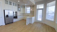 This single-story space showcases a well-appointed kitchen as its centerpiece. The kitchen is designed with crisp white shaker-style cabinetry that extends to the ceiling, providing generous storage throughout. Upper cabinets with glass-front doors add a refined touch, while a classic subway tile backsplash runs along the walls, complementing the light-toned countertops. A built-in wine rack adds a practical yet stylish accent above the counter.

Stainless steel appliances, including a double wall oven and built-in microwave, are seamlessly integrated into the cabinetry. A range hood in brushed steel adds a professional kitchen aesthetic. The peninsula, supported by elegant decorative columns, offers additional prep space and serves as a natural divider between the kitchen and the adjoining living area.

Pendant lights hang above the peninsula, providing focused task lighting. Natural light flows generously into the space through a large window above the sink and a glass-paneled exterior door, which opens to what appears to be a balcony or exterior walkway. Light hardwood flooring runs throughout, tying the space together with a warm, cohesive feel.
