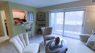 This single-floor space presents a thoughtfully arranged living area distinguished by sage green accent walls and light-toned carpeting that runs throughout. The living room serves as the central gathering area, furnished with cushioned seating and a curved glass-top coffee table that adds a contemporary touch. A tall, open shelving unit along one wall provides display space and contributes to the room's organized aesthetic.

A standout feature is the large sliding glass door fitted with vertical blinds, which floods the space with natural light and offers a view of lush outdoor greenery, creating a pleasant indoor-outdoor connection. The bright, airy atmosphere is enhanced by the neutral ceiling and light-colored carpet.

Adjacent to the living area, a breakfast bar with bar-height seating provides a seamless transition into the kitchen. The kitchen is visible in the background, featuring wood-toned cabinetry and a functional counter setup. The bar-style pass-through design encourages an open flow between the kitchen and living spaces, making the layout feel connected and practical for everyday living and casual entertaining.