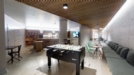 This contemporary single-floor amenity space is thoughtfully designed to blend recreation, socializing, and casual work in one cohesive environment. The space is anchored by a striking wood-slat ceiling that adds warmth and texture, complementing the polished concrete floors and neutral-toned walls for a clean, modern aesthetic.

At the center of the space sits a full-size foosball table, serving as a focal point for leisure and social interaction. Adjacent to it, a well-appointed bar area features dark wood cabinetry, open shelving stocked with glassware, and pendant lighting that creates an inviting atmosphere. Bar-height seating and countertops offer additional spots for informal gatherings or quick conversations.

On the opposite side of the space, a row of bright mint-green chairs lines communal tables, providing a lively and functional seating area ideal for casual meetings or collaborative work sessions. A wall-mounted television nearby adds to the space's versatility. Structural columns are seamlessly integrated into the open-plan layout, which benefits from generous ceiling height and well-distributed lighting throughout. The overall ambiance is energetic yet refined, making it a well-rounded space suited for both relaxation and productivity.
