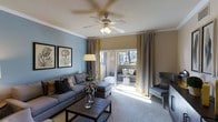 This single-story space centers around a thoughtfully designed living room that balances comfort and style. The room is finished with crown molding along the ceiling and features a ceiling fan with integrated lighting, contributing to both functionality and a polished aesthetic. Soft, neutral-toned carpet runs underfoot, complementing the cool blue-gray accent wall that adds depth and character to the space. Large sliding glass doors serve as a prominent architectural feature, flooding the interior with natural light and offering a direct view of — and access to — an outdoor patio. Floor-length curtains in gray with yellow accents frame the doors, adding a refined decorative touch. The living area is furnished with a spacious gray sectional sofa, an upholstered ottoman, and an accent chair, all arranged around a rustic wood coffee table. Botanical-themed wall art and a tall floor lamp contribute to the room's warm, curated atmosphere. The patio beyond the sliding doors appears to offer additional seating, extending the living space outdoors and enhancing the overall sense of openness and connectivity with the exterior environment.