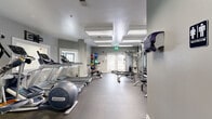 Located at 551 Settlers Landing Rd in Hampton, Virginia, this fitness center offers a well-equipped, single-floor workout facility designed to support a full range of exercise routines. The space is finished with cool gray walls, durable rubber flooring, and bright overhead fluorescent panel lighting that ensures a well-lit environment throughout.

The cardio section features multiple Precor treadmills and elliptical machines, providing ample options for aerobic training. A spinning bike adds to the cardiovascular offerings, and wall-mounted televisions are positioned for easy viewing during workouts. The strength training area is comprehensively outfitted with a TuffStuff Smith machine, a cable cross machine, multiple weight benches, and a full dumbbell rack. A medicine ball storage rack loaded with a colorful assortment of weighted balls supports functional fitness and core training.

Floor-to-ceiling mirrors line portions of the walls, visually expanding the space and allowing users to monitor their form. Natural light filters in through several windows and French doors, lending an open, airy feel to the interior. A built-in water fountain and sanitation station with cleaning supplies ensure cleanliness and convenience. The layout is thoughtfully organized, allowing for comfortable movement between equipment zones while maintaining a clean, professional fitness environment.