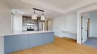 This contemporary single-floor residence showcases a thoughtfully designed open-concept layout with light hardwood flooring throughout the main living areas. The kitchen serves as the centerpiece of the space, anchored by a striking powder-blue island with a crisp white countertop and pendant lighting suspended from a dark wood bar above. Gray cabinetry lines the perimeter walls, complemented by a subway tile backsplash and a full suite of stainless steel appliances, including a gas range, built-in microwave, dishwasher, and a large French door refrigerator. A tall window at the end of the kitchen corridor draws in natural light and urban views.

The living area extends from the kitchen in an open, airy arrangement, featuring a built-in shelf along one wall. Bedrooms are carpeted and filled with natural light from large black-framed windows that frame panoramic city skyline views, including notable urban landmarks. A walk-in closet with built-in shelving and rod storage serves the primary bedroom.

Two bathrooms are finished with gray cabinetry and white countertops. The primary bath features a double-sink vanity with matte black fixtures, gold-toned lighting, and a wide frameless mirror, while the secondary bath offers a single-sink vanity with similar finishes.