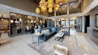 This upscale residential amenity lounge is a thoughtfully designed gathering space that blends sophisticated aesthetics with a warm, inviting atmosphere. The centerpiece of the main lounge is a soaring double-height ceiling adorned with an eclectic collection of sculptural pendant lights crafted from woven materials, casting a rich, ambient glow over the expansive seating area below. Large-format light wood flooring runs throughout, complemented by patterned area rugs with gold accents that anchor the various seating arrangements.

The lounge features plush sectional sofas and accent chairs arranged around coffee tables, encouraging relaxed conversation. A prominent wine display tower and dedicated wine bar area with shelving and signage serve as a focal point for entertaining. A separate game and entertainment zone includes a full-size billiards table with designer overhead lighting, wall-mounted flat-screen televisions, and a built-in wet bar with counter-height seating.

Floor-to-ceiling windows and glass doors flood the interior with natural light and offer views of the surrounding outdoor area. Decorative elements throughout — including driftwood sculptures, starburst wall art, and textured wall treatments in dark metal and wood — lend a curated, gallery-like quality to the space. A quieter lounge alcove with intimate booth-style seating provides an additional retreat within the amenity level.