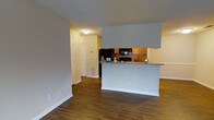 This single-floor residence is designed with an open-concept layout that promotes a sense of spaciousness and easy flow between living areas. The kitchen is a standout feature, positioned behind a raised peninsula counter that serves as both a functional workspace and a visual divider between the kitchen and the main living area. The kitchen is equipped with warm wood-toned cabinetry, dark appliances including a refrigerator and microwave, and a stone-look countertop that adds a touch of sophistication. Rich, dark hardwood-style flooring runs throughout the main living areas, providing a warm and cohesive aesthetic that contrasts elegantly with the light neutral-toned walls. The living area is generously proportioned, offering ample room for a variety of furniture arrangements. A hallway leads further into the residence, where a louvered closet door hints at convenient storage solutions. Subtle details such as clean white baseboards, a textured ceiling, and recessed floor vents complete the practical yet polished interior. The overall ambiance is bright, clean, and welcoming, making it a comfortable and versatile living space.