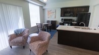 This single-floor unit is designed with an open-concept layout that seamlessly connects the living, dining, and kitchen areas into one cohesive space. The kitchen is outfitted with dark espresso cabinetry, black appliances including a gas range and built-in microwave, and a light-toned countertop island that doubles as a breakfast bar and prep surface. The kitchen flows naturally into the dining area, which features a compact round table with ladder-back chairs — a practical setup well-suited to everyday use. Adjacent to the dining space, the living area is furnished with patterned accent chairs arranged around a small side table, creating an inviting conversational nook. Hardwood-style flooring runs throughout, adding warmth and visual continuity. A large window floods the space with natural light, contributing to an open and airy atmosphere. Decorative touches, including a blue area rug and floral wall art, add personality. The overall design balances functionality with comfort in a well-organized, efficient floor plan.