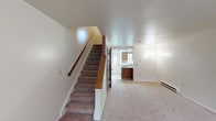 This two-story residence offers a practical and well-organized layout across both levels. The main floor welcomes you with a carpeted living area bathed in natural light, seamlessly connected to the kitchen. The kitchen is equipped with rich dark wood cabinetry extending to the upper walls, white countertops, a stainless steel sink, and a full suite of appliances including a range and dishwasher, all set on light tile flooring. A staircase with a warm wood handrail leads to the upper level, where the bedrooms are located. Each bedroom features plush carpeting, baseboard heating, and windows that invite natural light into the space. The upper floor also provides closet storage and easy access between rooms. The bathroom is appointed with a wood-framed vanity mirror, a single-basin sink set in a laminate countertop with wood cabinetry below, and durable wood-look vinyl flooring. Baseboard heaters are present throughout both floors, ensuring consistent warmth. The overall design is straightforward and functional, with neutral tones and classic wood accents creating a cohesive, comfortable atmosphere throughout the home.