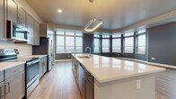 This contemporary single-floor residence showcases a thoughtfully designed open-concept layout with high-end finishes throughout. The centerpiece of the home is a sleek, modern kitchen featuring a generous waterfall-edge island with an undermount sink, crisp white quartz countertops, and gray shaker-style cabinetry reaching toward the ceiling. A suite of stainless steel appliances — including a French door refrigerator, range, and microwave — complements the textured tile backsplash. A sculptural oval pendant light anchors the space with stylish illumination. An expansive curved bay window wall floods the kitchen and living areas with natural light while framing sweeping panoramic views of the surrounding cityscape. The open living area continues the warm wood-look plank flooring and offers generous proportions with large windows further enhancing the bright, airy atmosphere. Bedrooms are carpeted for comfort and feature large windows with city views, along with a spacious walk-in closet equipped with built-in wire shelving. Two well-appointed bathrooms include vanities with white quartz countertops, gray shaker cabinetry with brushed nickel hardware, and wood-look tile flooring. One bathroom features a tiled walk-in shower, while the other offers a soaking tub.