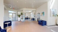 This contemporary amenity building is designed with a clean, modern aesthetic throughout its single floor. The welcoming lobby greets visitors with warm-toned flooring, recessed lighting, and stylish navy blue velvet seating that adds a bold pop of color against the crisp white walls. Tall, sculptural planters housing tropical palms serve as eye-catching focal points near the entrance, enhancing the space's inviting atmosphere.

A glass-walled office enclosure with aluminum framing sits prominently within the lobby, offering full transparency into the workspace within, which features seating, desks, and informational displays. An elevator and adjacent stairwell provide seamless vertical access, with lounge seating, colorful abstract artwork, and oversized windows framing exterior views positioned at key transition points throughout.

The fitness center is a standout feature, boasting high ceilings with exposed ductwork and industrial-style pendant lighting. Rows of cardio equipment — including treadmills, ellipticals, and stationary bikes — line one side, while a robust strength training area with a power rack, cable machines, weight benches, and a functional training tire occupy the opposite side. Large windows flood the space with natural light, creating an energizing environment well-suited for a comprehensive workout experience.