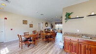 This single-floor community common area is designed to foster comfort and social interaction, seamlessly blending a dining space with a relaxing lounge area. The open-concept layout is unified by light wood-tone flooring that runs throughout, creating a warm and cohesive aesthetic.

The dining area features multiple wooden tables with cushioned chairs, accommodating a variety of group sizes. Large windows dressed with decorative valances allow ample natural light to fill the room, casting a bright and inviting atmosphere. Framed artwork adorns the walls, adding a homey touch to the space.

Transitioning naturally from the dining area, the lounge features rich brown leather seating, including a full sofa and accent chairs, arranged around a wooden coffee table. A decorative sunburst mirror serves as a focal point on the wall, complementing the warm tones of the furniture. A tall indoor plant adds a refreshing natural element nearby.

A wooden entertainment armoire housing a flat-screen television provides a media focal point in the lounge. Recessed lighting throughout ensures the space remains well-lit during all hours. A granite-topped cabinetry station near the entrance area offers a convenient service counter. The space is completed by a branded entryway mat, a wall clock, and thoughtful decorative greenery, all contributing to an organized, welcoming communal environment.