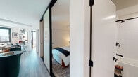 This single-floor residence showcases a thoughtfully designed layout that balances modern aesthetics with functional living. The open-concept kitchen and living area form the heart of the space, where the kitchen features striking two-tone cabinetry — crisp white upper cabinets contrasted by deep, dark lower cabinets — paired with stainless steel appliances and a clean subway tile backsplash. Light wood-tone flooring extends throughout, lending warmth and continuity across the living areas.

The living space benefits from expansive floor-to-ceiling windows framed in matte black, flooding the room with natural light and offering sweeping elevated city views. The primary bedroom is accessed through sleek black-framed glass sliding doors, providing a refined sense of separation while maintaining an open feel. It features a wall-mounted wood shelf, concrete accent column, and a sliding closet. A second bedroom mirrors similar contemporary styling, with large windows drawing in abundant sunlight and panoramic urban views, complemented by a built-in shelving and desk area. The bathroom is finished with large-format tile flooring, white tile surround, a soaking tub, and matte black fixtures throughout, maintaining the cohesive modern palette found across the entire residence.