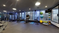 Located in Cleveland's vibrant East 12th Street corridor, this fitness center is a comprehensive, professionally appointed facility designed to support a full range of workout regimens. The space is divided into distinct functional zones, all unified by floor-to-ceiling windows that flood the interior with natural light and offer sweeping views of the Cleveland skyline.

The main strength training area is generously sized, housing a full complement of equipment including power racks, a Jones machine, cable systems, and an extensive dumbbell rack. Mirror-lined walls enhance the sense of depth throughout this zone. Adjacent to it, a dedicated cardio section features treadmills, ellipticals, and stationary bikes, all positioned to maximize the city views through the large-pane windows.

A functional training zone offers spin bikes, a Ski Erg machine, stability balls in multiple sizes, battle ropes, and a heavy tire, providing versatile options for high-intensity and cross-training workouts. Rubber flooring throughout the fitness areas ensures safety and durability.

Connected to the gym is a comfortable lounge space with hardwood-style flooring, a sectional sofa, a coffee table, and a dining area — ideal for relaxation before or after a workout. Recessed and panel lighting creates a welcoming ambiance throughout the entire facility.
