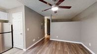 Located at 4250 E 29th St in Tucson, Arizona, this single-floor unit offers a functional and well-maintained layout. The living area is finished with warm wood-look vinyl plank flooring that runs throughout, complemented by neutral greige-toned walls and crisp white baseboards that create a clean, modern aesthetic. A ceiling fan with integrated lighting is centrally mounted on the textured ceiling, providing both airflow and illumination — an essential feature in the Tucson climate. A wall-mounted air conditioning unit near the entry ensures comfortable temperatures year-round. Natural light enters through a window adjacent to the front door, brightening the main living space. A dedicated closet provides storage, while a hallway leads to a bathroom featuring white cabinetry, a mirrored vanity with Hollywood-style lighting, and a light countertop — offering a polished, functional finish. The layout flows efficiently, with each area clearly defined despite the compact footprint. The consistent use of neutral tones, durable flooring, and practical fixtures throughout gives the space a cohesive and move-in-ready character, well-suited for comfortable everyday living in Tucson.