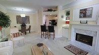 This single-floor residence offers a thoughtfully designed open-concept layout that seamlessly connects the main living areas. The living room serves as a central hub, anchored by an elegant white marble fireplace with a classic surround, adding a refined focal point to the space. Built-in shelving flanks the fireplace, providing display space for books and decorative accents. Warm neutral walls and plush wall-to-wall carpeting create a cohesive, comfortable ambiance throughout.

A raised breakfast bar with two dark wood bar stools transitions smoothly into the kitchen, which features white cabinetry and dark appliances visible through the pass-through opening. Trailing greenery along the upper cabinetry adds a natural, decorative touch. A dedicated dining area sits adjacent to the living space, illuminated by a pendant light and furnished with upholstered seating, ideal for gatherings.

A wet bar or butler's area with white cabinetry and under-cabinet lighting provides added functionality. The overall design balances classic elegance with practical living, making it a well-appointed and inviting space.