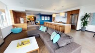 This single-floor space showcases a thoughtfully designed open-concept layout that seamlessly connects the living, kitchen, and dining areas into one cohesive and inviting environment. The living area is anchored by a generously sized gray sectional sofa accented with teal and turquoise throw pillows, complemented by a clean-lined white coffee table with yellow drawer accents — a subtle nod to the space's playful color palette.

The kitchen is a standout feature, centered around a substantial island with a crisp white countertop and warm wood cabinetry, lined with matching wooden bar stools. Pendant lights with natural wood shades hang above the island, reinforcing the earthy, contemporary aesthetic. Full-height cabinetry and a stainless steel refrigerator and range hood complete the well-appointed culinary space.

A bold blue accent wall frames a cozy dining nook in the background, adorned with framed artwork and built-in bench seating, creating an intimate contrast to the open main area. A warm wood feature wall anchors this zone, adding texture and depth. Recessed lighting throughout ensures the space is bright and welcoming. The overall ambiance is modern yet comfortable, balancing clean architectural lines with warm natural materials and lively pops of color.