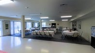 This single-floor community hall offers a generously sized, open gathering space designed to accommodate a variety of events and functions. The main hall is outfitted with multiple round tables draped in white linens and surrounded by cushioned chairs, providing comfortable seating for a large number of guests. Tasteful decorative accents, including floral centerpieces and heart-shaped wall ornaments, lend a warm and inviting ambiance to the room.

The space is well-lit by a series of recessed and overhead fluorescent light fixtures that ensure even illumination throughout. Large windows along one wall allow natural light to filter in, contributing to the bright and airy atmosphere. Structural support columns are positioned thoughtfully within the open floor plan, maintaining an unobstructed sense of spaciousness.

A flat-screen display mounted near the front of the room serves as a functional focal point, suitable for presentations or entertainment. The flooring transitions between light-toned hard surface and carpet, subtly defining different zones within the hall. A bulletin or display board is visible near the entrance area, suggesting the space is actively used for community programming. Overall, this hall is well-suited for banquets, community meetings, celebrations, and organized group activities.