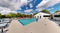 Located at 500 E Constitution Dr in Palatine, Illinois, this outdoor aquatic and recreation area offers a well-appointed community pool experience. The centerpiece is a generously sized rectangular swimming pool with clear, blue water, accessible via stainless steel entry rails and surrounded by a broad concrete sun deck. Rows of aqua-toned lounge chairs line the perimeter, providing ample space for sunbathing and relaxation. A hot tub is situated adjacent to the main pool, adding another option for leisure. The pool area is enclosed by a dark metal fence, ensuring safety while maintaining open sightlines. A white brick pool house anchors one side of the complex, featuring restroom facilities, posted pool rules, and large windows that connect interior and exterior spaces. A covered pergola area provides shaded seating for gatherings. The surrounding landscape is lush with mature trees, and the rear of the property offers a serene view of a tranquil pond, enhancing the natural ambiance of the setting. The overall design balances functional amenities with a comfortable, resort-inspired atmosphere suited for warm-weather recreation.