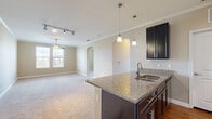 This single-floor residence offers a thoughtfully designed open-concept layout that seamlessly connects the kitchen and living area. The kitchen is a standout feature, boasting rich dark cabinetry, granite countertops, a central island with a built-in sink, elegant pendant lighting, and a full suite of stainless steel appliances including a refrigerator, electric range, and over-the-range microwave. Wood-look flooring grounds the kitchen space, while crown molding and track lighting add a polished finish. The open living area extends naturally from the kitchen, featuring soft carpeting, a ceiling fan, crown molding, and a window that invites natural light. An arched passageway adds an architectural accent that enhances the overall flow of the space. The bedroom is generously sized with plush carpeting, dual windows with blinds, and access to both a walk-in closet equipped with wire shelving and a full bathroom. The bathroom features a granite-topped dark vanity, wood-look flooring, and a shower-tub combination. Neutral warm tones are carried consistently throughout, creating a cohesive and inviting ambiance across every room.