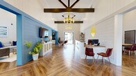 This single-story clubhouse is designed with a cohesive modern-farmhouse aesthetic, combining warm wood tones, shiplap wall treatments, and bold blue accent walls throughout. The entry reception area makes an immediate impression with soaring vaulted ceilings accented by exposed dark wood beams and a statement gold chandelier. Herringbone-patterned hardwood flooring flows seamlessly through the interior, anchoring the space with warmth and visual interest.

The reception desk, finished in crisp white, faces leather guest chairs and is flanked by a branded yellow logo accent wall. Adjacent to the entry, a lounge area features a bold blue shiplap accent wall with a mounted television and a console cabinet, opening to a casual seating area with a round dining table and floor-to-ceiling glass doors that invite natural light and direct views of the outdoor pool.

A dedicated conference room offers a long wood table, leather rolling chairs, a wall-mounted display screen, and large sheer-draped windows providing abundant natural light. The kitchenette is outfitted with white cabinetry, dark countertops, a beverage cooler, coffee station, and a striking blue subway tile backsplash.

Outdoors, a generous pool deck with lounge chairs, market umbrellas, and a covered cabana area overlooks the sparkling pool, completing this well-appointed amenity space.