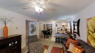 This single-story residence features two bedrooms and two bathrooms within a thoughtfully arranged floor plan. The open-concept living area serves as the heart of the home, where the living room, dining space, and kitchen flow naturally into one another, creating a connected and functional environment.

The living room is anchored by a ceiling fan with integrated lighting and features a textured ceiling, light-toned walls, and tile flooring. Decorative touches, including wall art and layered area rugs, add warmth to the space. A doorway from the living area leads toward the bedrooms and bathrooms, providing a clear separation between living and private spaces.

The dining area accommodates a full table and chair set, positioned adjacent to a built-in shelving unit that offers both storage and display space. The kitchen features a peninsula with bar-height seating for two, providing additional casual dining options. White cabinetry, a refrigerator, and a microwave are visible, along with warm wood-toned upper cabinetry that adds contrast. The layout allows for easy interaction between the kitchen and surrounding living and dining areas, making the space well-suited for everyday living and entertaining.