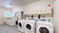 Located at 907 McAdoo Pl in Saanich, BC, this shared laundry facility is a practical, well-equipped space designed to serve multiple users efficiently. The room features a clean, utilitarian layout with white-painted walls, fluorescent overhead lighting, and a concrete floor that speaks to its durable, functional design.

The main area houses a row of commercial-grade front-load washing machines and top-load dryers, all finished in white and equipped with modern digital payment terminals for convenient use. A line of upper wall-mounted cabinets runs above the machines, providing ample storage space. Hose connections and utility hookups are neatly organized along the back wall.

A dedicated folding and sorting area is accessible via a side room, featuring a built-in countertop workspace along the wall, a cork bulletin board for notices, and a wall-mounted payment or access terminal. A fire extinguisher is also mounted nearby for safety compliance.

Toward the back of the room, a deep utility sink provides an additional washing station, accompanied by storage for cleaning equipment such as mops and buckets. A window allows a degree of natural light into the space. The facility is well-organized and designed to accommodate the laundry needs of a residential building with ease and practicality.