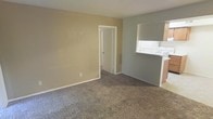 This single-floor residence features a modestly sized living area finished with wall-to-wall carpet in a neutral gray-brown tone, providing a comfortable and cohesive foundation throughout the space. The walls are painted in a warm beige, creating a simple, understated backdrop that lends itself well to a variety of interior styles.

A defining architectural feature of the living area is the pass-through opening on the right side of the room, which connects to the kitchen. This cutout allows for visual continuity between the two spaces and facilitates convenient interaction between the kitchen and living areas. The kitchen, partially visible through the pass-through, includes upper cabinetry in a light wood tone and what appears to be standard countertop appliances or fixtures.

An interior doorway on the far wall of the living area provides access to additional rooms within the unit, suggesting a straightforward and functional layout. Standard white baseboards frame the carpeted floor throughout, adding a clean, finished look to the space. Overall, the residence offers a practical, no-frills environment with a neutral palette that provides a solid canvas for personalization.