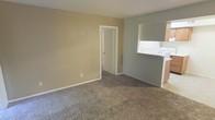 This single-floor residence features a modestly sized living area finished with wall-to-wall carpet in a neutral gray-brown tone, providing a comfortable and cohesive foundation throughout the space. The walls are painted in a warm beige, creating a simple, understated backdrop that lends itself well to a variety of interior styles.

A defining architectural feature of the living area is the pass-through opening on the right side of the room, which connects to the kitchen. This cutout allows for visual continuity between the two spaces and facilitates convenient interaction between the kitchen and living areas. The kitchen, partially visible through the pass-through, includes upper cabinetry in a light wood tone and what appears to be standard countertop appliances or fixtures.

An interior doorway on the far wall of the living area provides access to additional rooms within the unit, suggesting a straightforward and functional layout. Standard white baseboards frame the carpeted floor throughout, adding a clean, finished look to the space. Overall, the residence offers a practical, no-frills environment with a neutral palette that provides a solid canvas for personalization.