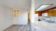 This single-floor residence offers a thoughtfully designed layout that balances comfort and functionality. The kitchen stands out with warm wood cabinetry, granite-style countertops, and a peninsula bar with seating for two — ideal for casual dining. A skylight panel above provides bright, even illumination throughout the cooking area, which is equipped with white appliances including a refrigerator, range, and microwave. The kitchen opens toward the living area, creating a connected and social floor plan.

The living space features light gray walls, textured ceilings, and updated wood-look vinyl flooring that runs consistently throughout the unit, lending a cohesive and modern feel. Plantation shutters on the windows offer both privacy and control over natural light. The bedroom is generously sized and includes a ceiling fan with light fixture and a full-width mirrored sliding closet, maximizing both storage and the sense of space. A smaller secondary room with its own plantation-shuttered window adds versatility as an office or additional bedroom.

The bathroom is clean and functional, featuring a full tub and shower combination with a curved rod and multi-function showerhead, complemented by chrome fixtures, a granite-topped oak vanity, and wood-look flooring that ties the space together seamlessly.