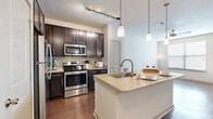 This thoughtfully designed one-bedroom, one-bathroom residence offers a functional and stylish single-floor layout. The kitchen serves as the heart of the home, showcasing rich dark wood cabinetry, a mosaic tile backsplash, and a full suite of stainless steel appliances including a range, microwave, and refrigerator. A central island with a granite countertop and built-in sink provides additional prep space and casual dining, illuminated by elegant pendant lighting overhead. Track lighting further enhances the kitchen's bright and inviting atmosphere. Warm hardwood-style flooring extends throughout the main living and kitchen areas, creating a cohesive and polished look. The bedroom is a comfortable retreat, featuring plush neutral carpeting, a large window that invites natural light, and a ceiling fan for year-round comfort. A dedicated closet offers convenient storage. The bathroom is finished with matching granite countertops, dark-framed cabinetry, a large mirror, and a full tub and shower combination tiled in classic white subway tile, complemented by the same hardwood-style flooring. The overall ambiance is modern and clean, with a neutral color palette that creates a warm, welcoming environment throughout.
