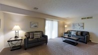 This single-floor space offers a welcoming and functional layout designed for comfortable everyday living. The living area serves as the centerpiece of the home, featuring wall-to-wall carpeting that adds warmth underfoot and a soft, neutral palette throughout. A prominent set of floor-to-ceiling sliding glass doors dressed with vertical blinds floods the room with natural light and provides a seamless connection to the outdoors. The textured ceiling adds a subtle architectural detail, while the neutral beige walls create a versatile backdrop that complements a variety of décor styles. The living room offers generous space for seating arrangements, as evidenced by the ample room for a sofa, accent chair, and coffee table. Framed artwork adorns the walls, enhancing the overall sense of a curated, lived-in atmosphere. The space is well-ventilated with ceiling vents ensuring year-round climate comfort. Overall, the space exudes a relaxed, approachable ambiance — practical in its design while remaining inviting and cozy, making it well-suited for both everyday relaxation and casual entertaining.