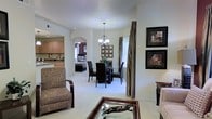 This single-floor residence offers an open-concept floor plan that effortlessly connects the main living areas, creating a cohesive and welcoming environment. The living room provides a comfortable gathering space, featuring plush seating, a glass-topped coffee table, and warm neutral carpeting that extends throughout the home. Framed artwork adorns the walls, adding a polished, decorative touch to the space.

The dining area is positioned centrally and anchored by a round glass-top table surrounded by dark wood chairs, all illuminated by an elegant chandelier that serves as a focal point overhead. Decorative wall art and tall draping curtains frame a bright window, allowing natural light to filter into the room.

A graceful arched passageway connects the main living and dining spaces to the kitchen, which features warm wood cabinetry and a functional layout. Through the archway, a glimpse of a bedroom is also visible, suggesting a well-organized and flowing floor plan.

The consistent use of light-toned walls and carpeting throughout creates a bright, airy ambiance, while the cohesive décor choices lend the space a refined and comfortable character. Overall, this residence offers a practical yet stylish single-level living experience.