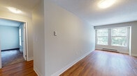 Located at 3–9 Upper River Road in South Hadley, Massachusetts, this single-floor unit offers a practical and comfortable layout with a clean, neutral aesthetic throughout. The living area features warm hardwood flooring, light gray walls, and a textured ceiling, with a large window that brings in natural light and views of surrounding greenery. A baseboard heating system runs along the perimeter, ensuring year-round comfort. The kitchen is equipped with white cabinetry, granite countertops, a coil-top electric range with an overhead hood, and ample storage space. The bedrooms offer a quieter atmosphere with plush carpeting, neutral-toned walls, and windows fitted with blinds overlooking outdoor foliage. One bedroom includes a generous closet with a rod and shelving. The bathroom features a white vanity with cabinet storage, chrome hardware including a towel bar and toilet paper holder, and a full tub-and-shower combination with a warm-toned tile surround. The overall layout is efficient and well-organized, with hardwood flooring connecting the main living spaces and a hall leading to the private areas of the unit.