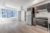 Located at 50 Portland Street in Toronto's vibrant King West neighbourhood, this one-bedroom, one-bathroom unit offers a well-designed layout across a single floor. Light-toned hardwood flooring runs throughout, creating a clean, cohesive aesthetic that complements the neutral colour palette.

Entry is through a bright hallway that leads into the open living and kitchen area. The kitchen is outfitted with dark cabinetry, a white subway tile backsplash, stainless steel appliances including a refrigerator and stove, and a dishwasher — offering a modern, functional workspace.

The bedroom is a well-proportioned space bathed in natural light from large floor-to-ceiling windows that frame views of the surrounding Toronto cityscape. A built-in double closet provides ample storage, and a stacked in-unit washer and dryer is conveniently tucked into a dedicated nook within the room.

The bathroom features light wood flooring, a white vanity with an illuminated mirror, and a full soaker tub with marble-style tile surround, lending a clean and polished finish. Throughout the unit, recessed and flush-mount lighting, white baseboards, and neutral walls contribute to a bright, move-in-ready atmosphere well-suited to urban living.