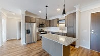 This single-floor residence showcases a thoughtfully designed open layout connecting the main living and kitchen areas. The kitchen serves as a central highlight, featuring rich, medium-toned shaker-style cabinetry, granite countertops with subtle veining, and a full suite of stainless steel appliances including a French door refrigerator, electric range, built-in microwave, and dishwasher. A generous island with matching granite provides additional prep space and casual seating. A herringbone-patterned tile backsplash behind the sink adds a refined decorative touch, while two sleek black cone pendant lights illuminate the workspace below.

Light wood-tone luxury vinyl plank flooring flows throughout the main living areas, complementing the neutral gray wall palette and crisp white crown molding. The living area is bright and open, benefiting from natural light through a sliding glass door that leads to a private balcony. The entry foyer transitions seamlessly into the kitchen and living space.

The primary bathroom includes a standalone walk-in glass shower, a vanity with granite countertop and dark cabinetry, and direct access to a walk-in closet equipped with wire shelving systems. A secondary bathroom features a full tub-shower combination with clean white surround finishes.