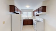 This single-floor unit offers a well-organized layout that maximizes functionality and livability. The interior is anchored by warm, richly toned hardwood floors that extend throughout the living and bedroom areas, lending a cohesive and inviting character to the space.

The kitchen is designed in a classic U-shape, featuring deep-toned dark wood cabinetry that provides generous storage both above and below the counters. White appliances — including a gas range with overhead ventilation hood and a full-size refrigerator — are neatly integrated into the layout. A single-basin sink is positioned beneath a double window, allowing natural light to brighten the workspace.

The living areas are open and airy, with clean white walls and multiple windows fitted with blinds that allow for adjustable natural light. Baseboard heating units are present throughout, ensuring consistent warmth. The rooms offer ample space for a variety of furniture arrangements.

The bathroom is finished in a clean, minimal style, featuring a full-size soaking tub with a shower fixture, neutral tile surround, and a window that provides natural ventilation and light.

Overall, the space presents a straightforward and practical floor plan with classic finishes, well-maintained hardwood flooring, and sufficient natural light throughout.
