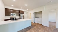 This single-floor unit is designed with a clean, contemporary aesthetic that prioritizes both functionality and comfort. The open-concept layout connects the kitchen and living area seamlessly, creating a spacious, airy feel enhanced by recessed lighting and crisp white walls throughout.

The kitchen is a standout feature, equipped with dark espresso cabinetry, stainless steel appliances including a refrigerator, range, and microwave, and a white quartz peninsula that provides ample counter space and serves as a natural divider between the kitchen and living area. An integrated sink with a single-handle faucet adds a streamlined touch.

The living area is bright and inviting, anchored by a large picture window that frames views of surrounding evergreen trees and fills the space with natural light. A wall-mounted HVAC unit ensures year-round climate control.

A dedicated in-unit laundry alcove houses a full-size washer and dryer alongside built-in shelving, offering practical storage. The bathroom features a dark wood vanity with white quartz countertop, a large mirror, and a soaking tub, with direct access to an adjacent walk-in closet equipped with built-in shelving and hanging rods. Hardwood-style flooring flows consistently throughout the entire unit.