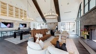 This single-story space showcases a striking contemporary design that blends modern and rustic elements throughout its open, expansive layout. The main gathering area commands attention with soaring vaulted ceilings accented by bold, dark exposed beams and two large circular chandelier fixtures with Edison-style bulbs, creating a warm yet sophisticated ambiance. Dark hardwood flooring runs throughout, grounding the bright white walls and ceiling.

A dramatic floor-to-ceiling dark stone fireplace serves as a central focal point, flanked by expansive black-framed glass windows and doors that invite generous natural light and offer views of the surrounding outdoor landscape. The lounge area is furnished with a mix of cream and cognac leather seating arranged around a wooden coffee table, providing a comfortable and inviting atmosphere.

A dedicated entertainment zone features a pool table, wall-mounted televisions set against a textured wood accent wall, and a bar counter with seating. Adjacent seating nooks with floor-to-ceiling windows offer quieter retreat spaces with outdoor views. A separate glass-enclosed room visible from the main area appears to house additional recreational space, such as a ping pong table, ensuring a variety of leisure options throughout.