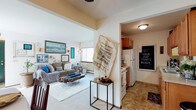 This single-floor unit is designed with an open, flowing layout that connects the living area and kitchen through a wide passthrough opening, creating a cohesive and functional living environment. The living room features wall-to-wall carpeting, white walls, and a large window that allows natural light to filter into the space. A baseboard heating unit runs along the exterior wall beneath the window, indicating an efficient heating setup. The room is comfortably sized, offering enough space for a full seating arrangement centered around a distinctive live-edge wood coffee table atop a plush area rug. Decorative wall art in coastal blue and neutral tones adds character to the space. Adjacent to the living area, the kitchen is finished with tile flooring, warm wood cabinetry, and white appliances including a refrigerator and range. The kitchen is compact yet functional, with sufficient counter space and storage. A ceiling fixture provides overhead lighting. The transition between the two spaces is framed by a clean archway, maintaining separation while keeping the overall layout open and connected. The unit presents a cozy, livable atmosphere throughout.