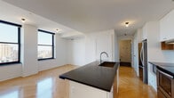 Located on the 18th floor of a modern high-rise in the heart of downtown Detroit, this unit at 1150 Griswold St offers a well-designed open-concept layout with contemporary finishes throughout. The herringbone-patterned hardwood flooring runs seamlessly across the entire unit, lending warmth and visual interest to every room.

The kitchen serves as a central hub of the space, anchored by a large island featuring a dark stone countertop and an integrated sink with a sleek stainless steel faucet. White shaker cabinetry lines the walls, complemented by warm wood open shelving and a classic white subway tile backsplash. Stainless steel appliances, including a full-size range, microwave, and refrigerator, complete the culinary space.

The open living area flows naturally from the kitchen, bathed in natural light from large windows that frame sweeping views of the Detroit skyline. The bedroom continues this connection to the city with corner windows offering elevated, panoramic views of the surrounding urban landscape.

The bathroom is finished with dark grid mosaic floor tiles, a wood-toned vanity with a white countertop, and black tile accents in the shower, reflecting a cohesive modern aesthetic. Gold hardware details throughout add a refined touch to the overall design of the unit.