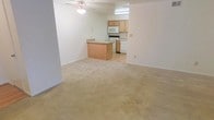 This single-floor space showcases an open-concept layout that seamlessly connects the living area to the kitchen. The living area is finished with plush, neutral-toned carpeting that extends throughout, creating a warm and cohesive feel. A ceiling fan is centrally positioned overhead, providing both airflow and a subtle design element.

The kitchen transitions from the carpeted living space via a breakfast bar peninsula, which offers additional seating and counter space while maintaining the open flow between areas. The kitchen itself features light wood cabinetry with ample storage, a white electric range, a built-in microwave, and a dishwasher. The kitchen floor is finished with smooth hard flooring, offering a practical contrast to the carpeted living space.

The walls throughout are painted in a clean, neutral white, lending brightness to the interior. Recessed under-cabinet lighting in the kitchen adds functional illumination to the work area. The overall design is straightforward and functional, with a neutral palette that provides a versatile canvas adaptable to a variety of furnishing styles and personal preferences.