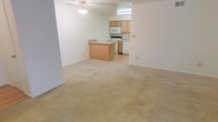 This single-floor space showcases an open-concept layout that seamlessly connects the living area to the kitchen. The living area is finished with plush, neutral-toned carpeting that extends throughout, creating a warm and cohesive feel. A ceiling fan is centrally positioned overhead, providing both airflow and a subtle design element.

The kitchen transitions from the carpeted living space via a breakfast bar peninsula, which offers additional seating and counter space while maintaining the open flow between areas. The kitchen itself features light wood cabinetry with ample storage, a white electric range, a built-in microwave, and a dishwasher. The kitchen floor is finished with smooth hard flooring, offering a practical contrast to the carpeted living space.

The walls throughout are painted in a clean, neutral white, lending brightness to the interior. Recessed under-cabinet lighting in the kitchen adds functional illumination to the work area. The overall design is straightforward and functional, with a neutral palette that provides a versatile canvas adaptable to a variety of furnishing styles and personal preferences.