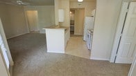 This single-floor unit features an open layout that seamlessly connects the kitchen and living area. The kitchen is equipped with white cabinetry, a white electric range, and a full-size refrigerator. A peninsula with a countertop overhang serves as a dividing element between the kitchen and the carpeted living space, offering additional prep space and a casual dining option. Light tile flooring runs through the kitchen area, transitioning to wall-to-wall carpet in the living room, creating a clear delineation between the two zones. Crown molding along the upper walls adds a subtle architectural detail. A hallway visible from the kitchen leads further into the unit, hinting at additional rooms beyond. The overall color palette is neutral, with beige and white tones throughout, contributing to a clean and functional aesthetic. The space appears well-maintained and offers a practical layout suited for everyday living.