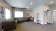 This single-floor space offers a clean, functional layout designed for comfort and practicality. The main lounge area is anchored by a generously sized gray sectional sofa, providing ample seating and a welcoming atmosphere. Recessed lighting in the smooth ceiling illuminates the room evenly, complementing the natural light that filters in through a window fitted with a white blind.

The walls feature a two-tone color scheme, with a muted gray-purple accent wall contrasting against crisp white walls, lending the space a modern yet understated aesthetic. Neutral gray carpeting runs throughout, adding warmth and cohesion to the interior.

Along one wall, a built-in workstation area includes wooden cabinetry and counter space, accompanied by a tall storage cabinet, making the space versatile for both relaxation and light task work. A water dispenser is conveniently positioned nearby. A small decorative side table rounds out the furnishings, adding a subtle design touch.

The overall ambiance is calm and orderly, with a straightforward layout that maximizes usable floor space. The combination of practical storage solutions, comfortable seating, and balanced lighting makes this a well-suited environment for a lounge, waiting area, or multipurpose room.