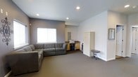 This single-floor space offers a clean, functional layout designed for comfort and practicality. The main lounge area is anchored by a generously sized gray sectional sofa, providing ample seating and a welcoming atmosphere. Recessed lighting in the smooth ceiling illuminates the room evenly, complementing the natural light that filters in through a window fitted with a white blind.

The walls feature a two-tone color scheme, with a muted gray-purple accent wall contrasting against crisp white walls, lending the space a modern yet understated aesthetic. Neutral gray carpeting runs throughout, adding warmth and cohesion to the interior.

Along one wall, a built-in workstation area includes wooden cabinetry and counter space, accompanied by a tall storage cabinet, making the space versatile for both relaxation and light task work. A water dispenser is conveniently positioned nearby. A small decorative side table rounds out the furnishings, adding a subtle design touch.

The overall ambiance is calm and orderly, with a straightforward layout that maximizes usable floor space. The combination of practical storage solutions, comfortable seating, and balanced lighting makes this a well-suited environment for a lounge, waiting area, or multipurpose room.