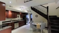 This two-story residence offers a thoughtfully designed main level that seamlessly integrates the kitchen and dining area into a cohesive, open living space. The kitchen is outfitted with rich, dark wood cabinetry paired with granite countertops and a full suite of stainless steel appliances, including a French-door refrigerator and dishwasher. Track lighting mounted on the ceiling provides targeted illumination throughout the kitchen workspace, enhancing the room's functionality and ambiance.

Adjacent to the kitchen, the dining area accommodates a round white table surrounded by patterned upholstered chairs, creating an inviting space for meals. The large-format white tile flooring extends throughout the main level, lending a clean and cohesive aesthetic to the open layout.

A standout architectural feature is the staircase, which showcases a dark wood frame accented by ornamental wrought iron balusters with a decorative spindle design. This staircase serves as both a functional and visual focal point, connecting the main level to the upper floor. The neutral white walls and bright lighting contribute to an overall atmosphere that feels open, airy, and well-appointed.