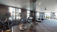 This contemporary fitness center is designed to support a full range of workout routines in a sleek, modern environment. The space is finished with a distinctive checkered tile-patterned wall treatment in shades of gray, lending a sophisticated aesthetic throughout. Durable rubber flooring in a complementary checkerboard pattern provides both traction and comfort underfoot.

The cardio zone is lined with large windows offering views of the surrounding urban streetscape, keeping the area bright and energizing. Equipment includes elliptical trainers, treadmills, stationary bikes, and rowing machines, thoughtfully arranged to maximize the open floor plan. Mirror-clad columns and full wall mirrors visually expand the space while allowing users to monitor their form.

The strength training area is equally well-appointed, featuring a complete rack of dumbbells, a cable crossover machine, a multi-functional weight rack with resistance training accessories, and a dedicated bench press station. A separate functional training zone offers medicine balls, TRX suspension straps, and additional resistance tools mounted on a storage rack. Recessed lighting and ceiling fans ensure the environment remains comfortable throughout a workout. The overall ambiance is clean, modern, and professionally outfitted, making this fitness center a standout amenity.