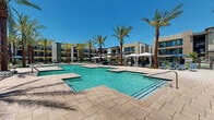 Located at 15400 W Roosevelt St in Goodyear, Arizona, this outdoor amenity space delivers a resort-caliber experience within a modern residential community. The centerpiece is a generously sized, multi-section swimming pool with crystal-clear turquoise water, framed by wide stone-paved pool decks that provide ample space for lounging and socializing. Towering palm trees are planted throughout the courtyard, offering natural shade and a distinct Southwestern aesthetic that complements the bright Arizona sunshine and vivid blue skies above.

A dedicated spa with seating areas is positioned adjacent to the main pool, offering a more relaxed retreat. The deck is outfitted with navy blue chaise lounges, side tables, and shaded umbrella stations, while private cabanas with flowing white curtains line one side of the pool for more secluded relaxation. Wrought iron fencing with gated access encloses the pool area, ensuring privacy and security for residents.

The surrounding multi-story residential buildings feature contemporary architecture with clean lines, balconies, and large windows that overlook the courtyard. Manicured green landscaping and decorative ground-level plantings add color and texture throughout the space, creating a cohesive and inviting outdoor environment well-suited to the warm desert climate of the greater Phoenix area.