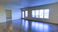 This single-floor space offers a clean, modern layout designed to maximize comfort and functionality. The main living area is immediately striking, featuring rich, dark hardwood floors that reflect the abundant natural light pouring in through a series of large windows fitted with horizontal blinds. The windows span a generous portion of the exterior wall, ensuring the interior remains bright and airy throughout the day.

A glass-paneled door provides direct access to an outdoor balcony, seamlessly connecting the interior living space to the outside. The neutral, light-toned walls create a versatile backdrop that complements a wide range of interior design styles, while the smooth, well-finished ceiling adds to the sense of openness and cleanliness throughout.

The open floor plan allows for flexible furniture arrangements, making it easy to define distinct living zones within the space. Recessed lighting in the ceiling supplements the natural light for evening use. The overall ambiance is bright, airy, and inviting, with a contemporary feel underscored by the quality of the flooring and the thoughtful placement of windows. This space presents a well-maintained, move-in-ready environment ideal for those seeking a modern, light-filled living experience.