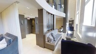 This contemporary lobby showcases refined architectural design with an emphasis on clean lines and upscale finishes. The space commands attention with its soaring double-height ceilings, enhanced by a dramatic curved white architectural beam that sweeps across the upper level, adding visual dynamism to the interior.

A standout decorative panel — featuring an intricate dark textured design framed in chrome hardware — serves as a central focal point, mounted against a taupe-toned structural column. Built-in bench seating with plush grey upholstery and coordinating throw pillows is integrated directly into the base of the column, offering a functional yet stylish resting area.

The flooring throughout is a smooth, light-toned polished surface that reflects the recessed ceiling lighting above, contributing to a bright and airy atmosphere. Elevator doors finished in a warm grey tone are visible along the corridor, suggesting this is a shared building lobby. Additional seating, including a deep navy blue chair and side table, further enhances the lounge-like quality of the space. Natural light filters in from glazed upper-level panels, completing the elegant, hotel-inspired ambiance.